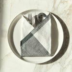 Image of M+A NYC Colorblock Napkin in Kora/Charcoal folded on a cream plate with a set of flatware tucked inside.