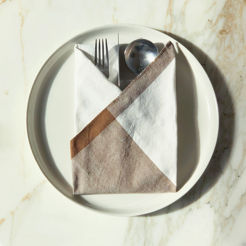Image of M+A NYC Colorblock Napkin in Kora/Earth folded on a cream plate with a set of flatware tucked inside.
