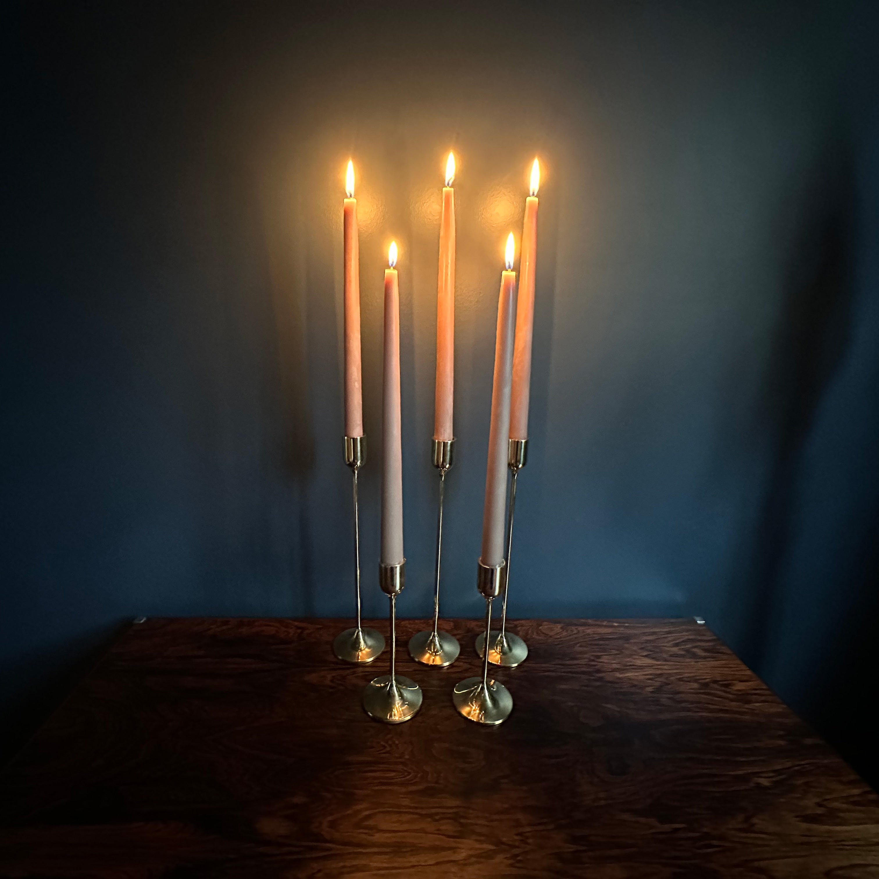 Image of 5 M+A NYC Solid Brass Taper Stands (3 Tall and 2 Small) with lit candles sitting atop a rosewood table against a blue green backdrop.