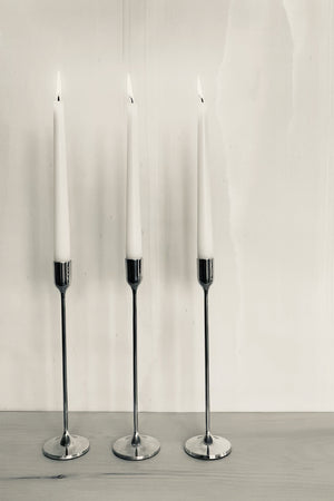 Image of 3 Tall Solid Brass Taper Stands with lit candles on a wooden mantel against pale iridescent silk wallpaper by Fromental. Shown with a silvertone filter.