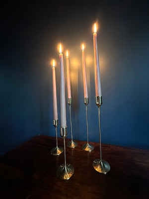 Image of 5 M+A NYC Solid Brass Taper Stands (3 Tall and 2 Small) with lit candles sitting atop a rosewood table against a blue green backdrop.