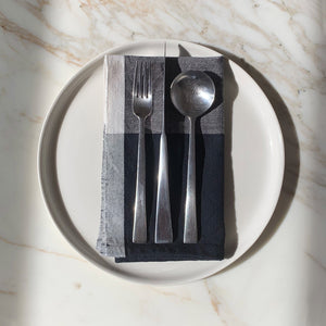 Image of M+A NYC Colorblock Napkin in Mineral/Kora folded on a cream plate with a set of flatware placed on top.
