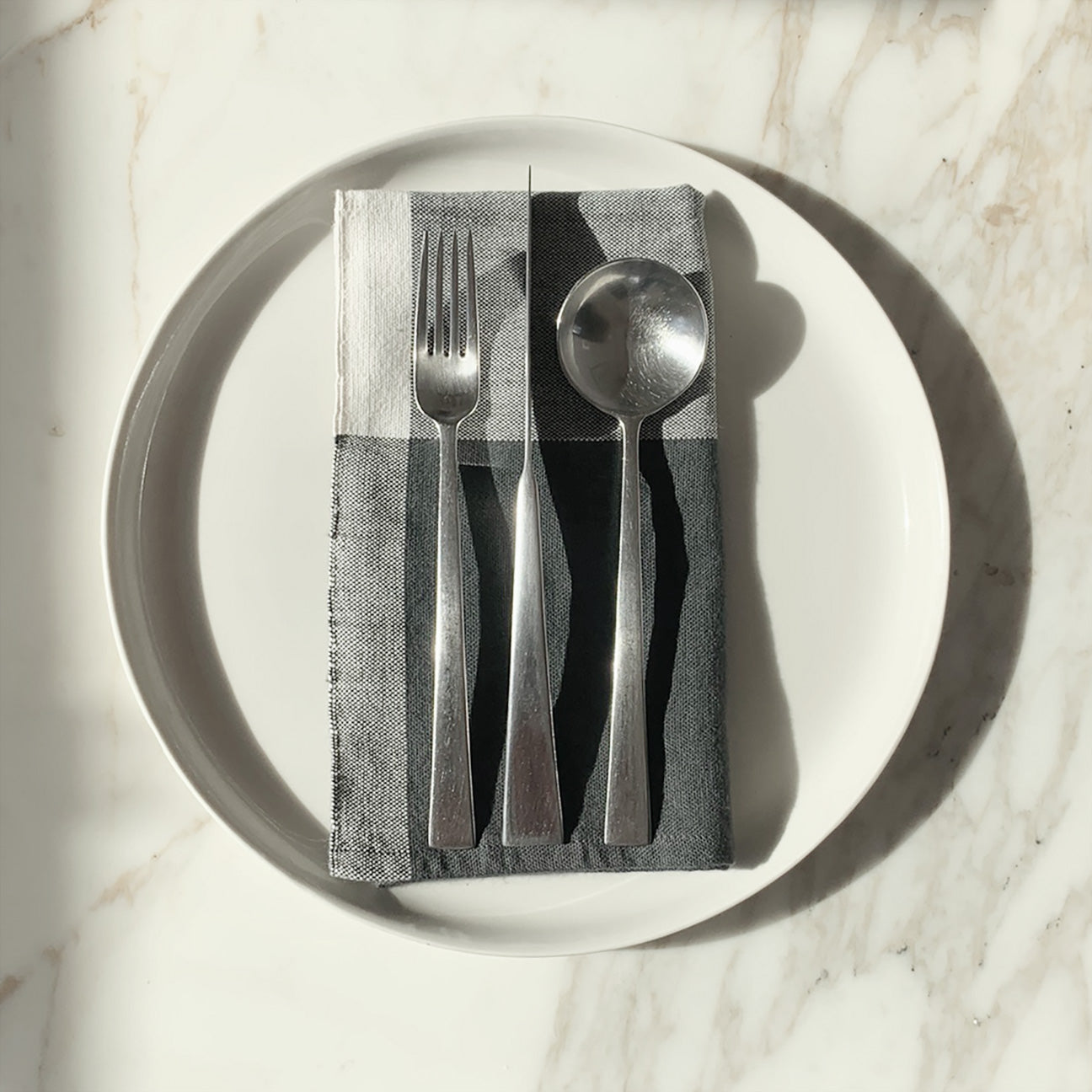 Image of M+A NYC Colorblock Napkin in Charcoal/Kora folded on a cream plate with a set of flatware placed on top.