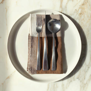 Image of M+A NYC Colorblock Napkin in Earth/Kora folded on a cream plate with a set of flatware placed on top.