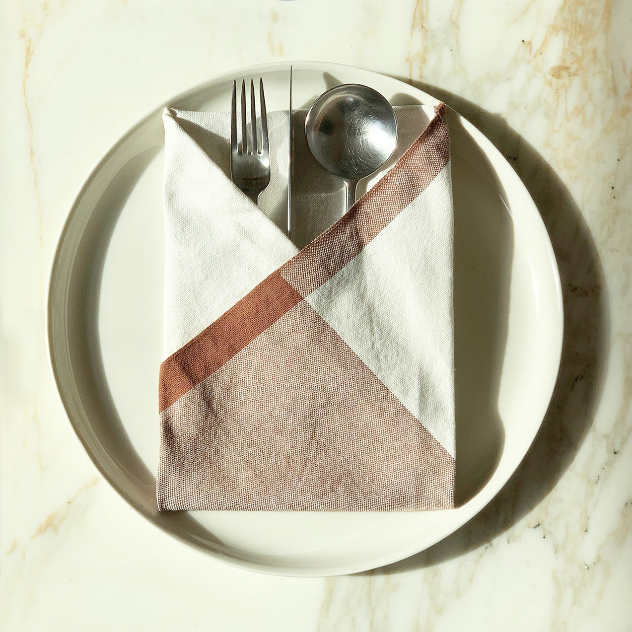 Image of M+A NYC Colorblock Napkin in Kora/Cafe Con Leche folded on a cream plate with a set of flatware tucked inside.
