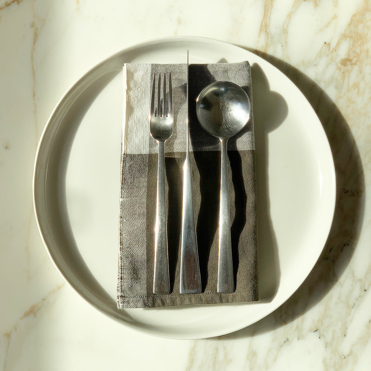 Image of M+A NYC Colorblock Napkin in Mineral/Kora folded on a cream plate with a set of flatware placed on top.