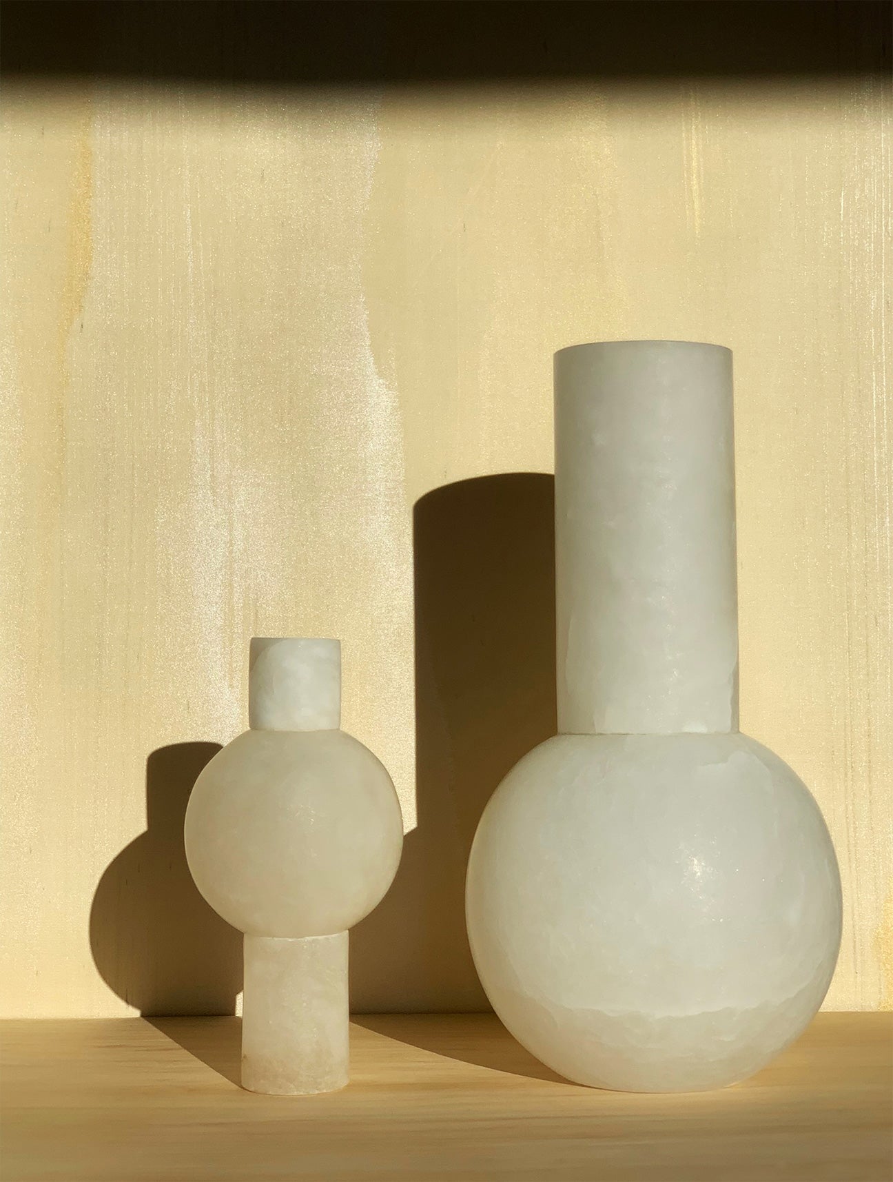 Image of M+A Keyhole Vase next to Lolita Vase, both in alabaster.  Sitting in front of wallpaper by Fromental Design in the pattern "Travertine".